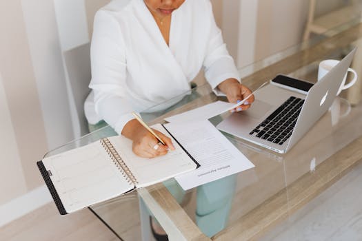 pexels-photo-5387256-5387256 Woman writing notes while working with a laptop and documents at a glass desk indoors.